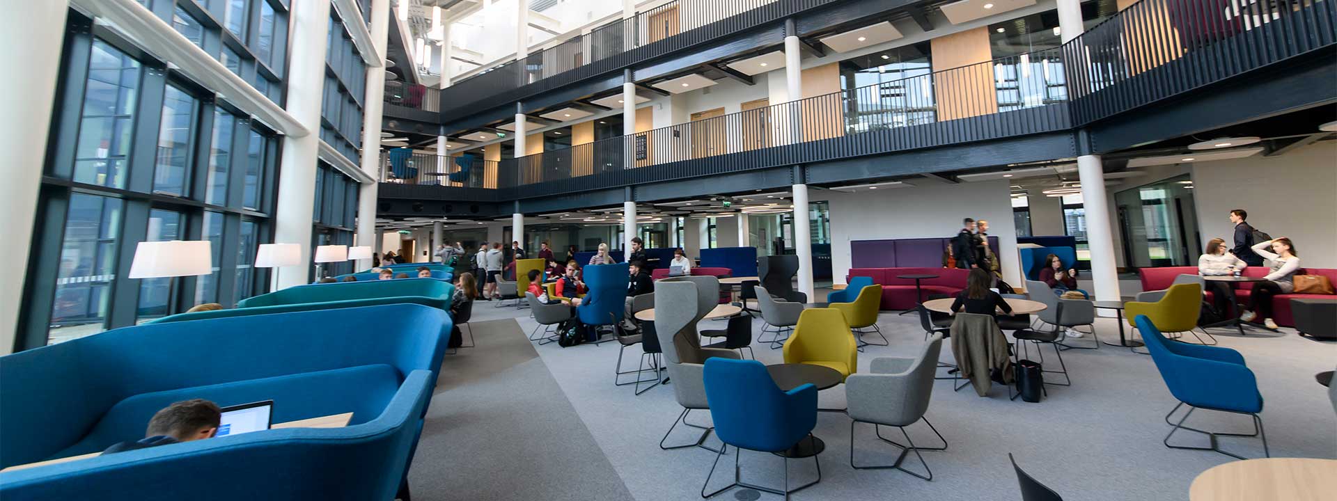 Modern university atrium with students seated in colorful chairs and booths, studying and socializing in a bright, open space with large floor-to-ceiling windows and multiple levels.