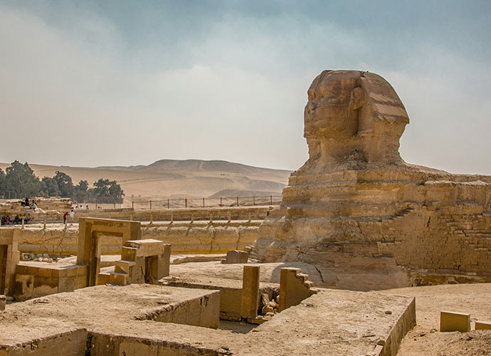 The Great Sphinx of Giza in Egypt, a colossal limestone statue with a lion’s body and a human head, surrounded by ancient ruins under a hazy sky with desert hills in the background.
