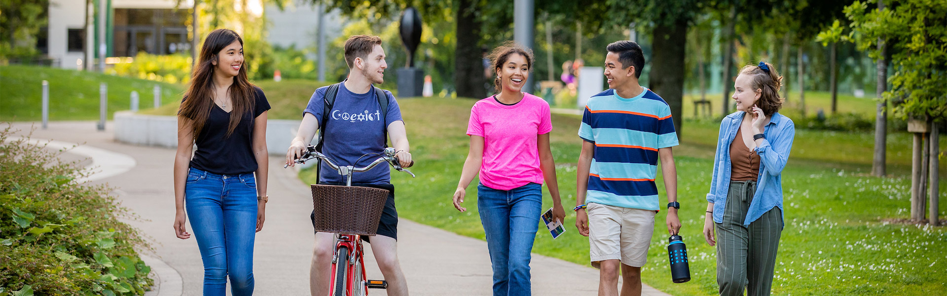 Five students walk together in a grassy campus. One has a bicycle.