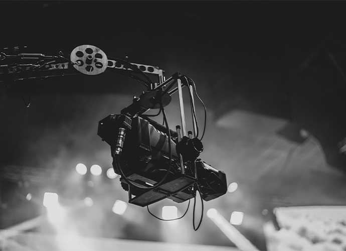 A professional video camera mounted on a robotic crane arm, capturing footage on a stage with bright spotlights in the background, in black and white.