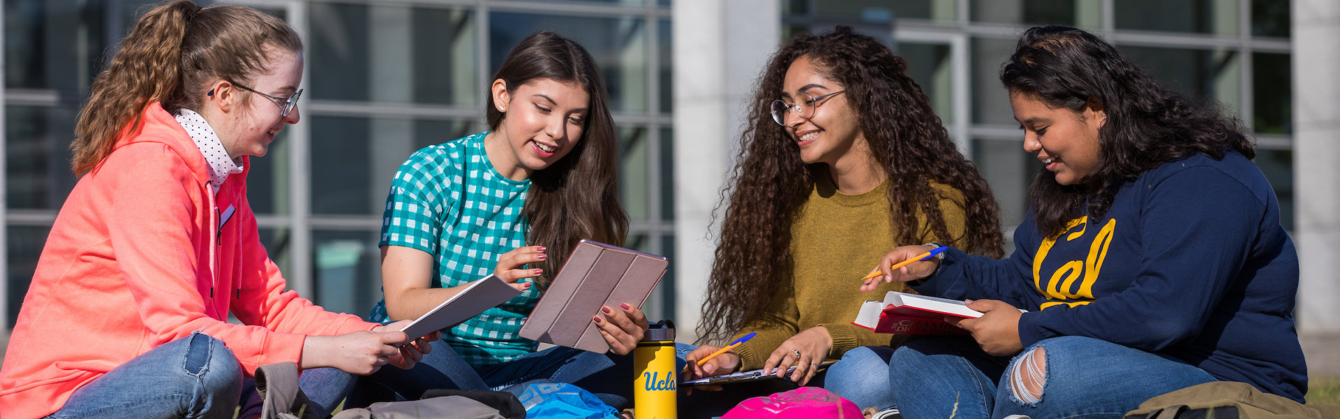 Four students sit on the lawn surrounded by books and bags.