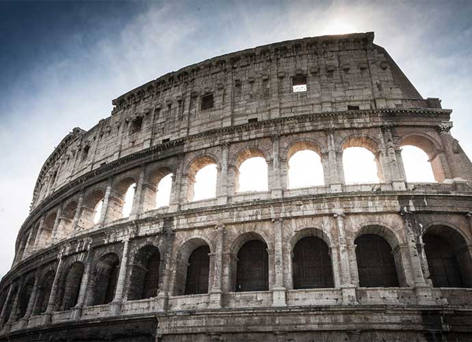 Exterior view of the Colosseum in Rome, showing its ancient stone arches and partial upper structure against a cloudy sky.