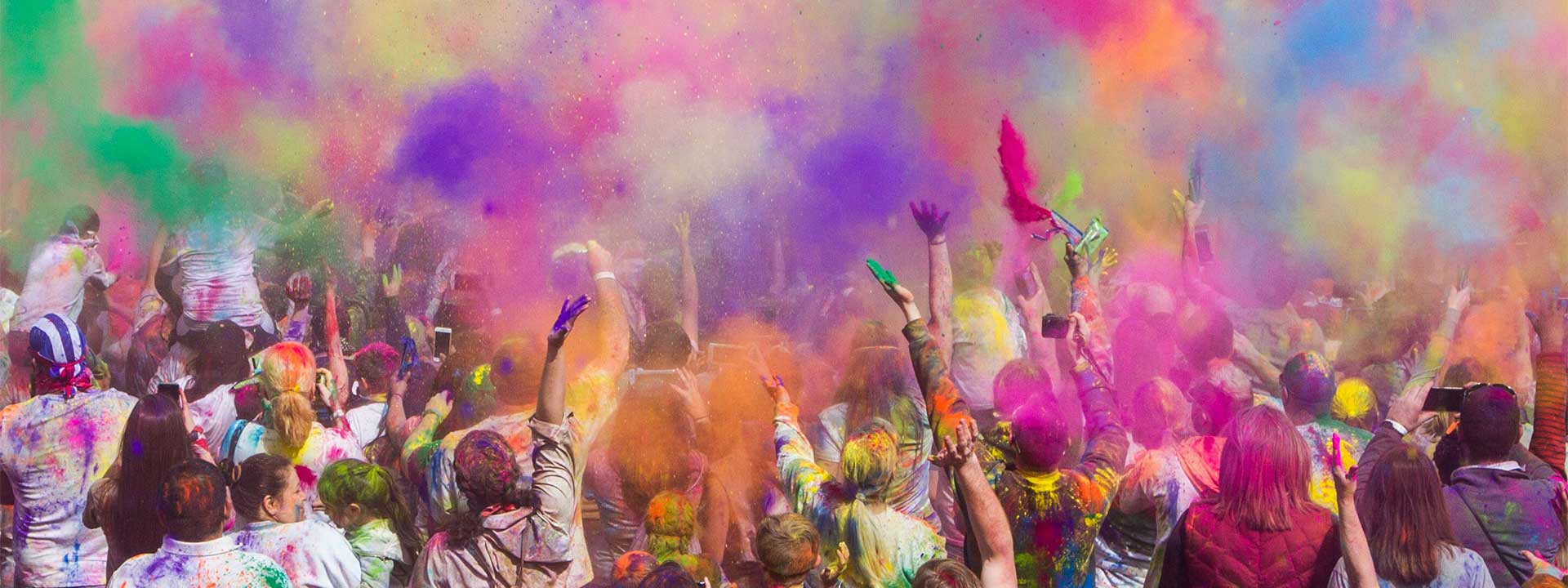 A large crowd of people celebrating Holi, throwing vibrant colored powders into the air, creating a cloud of pink, purple, green, yellow, and blue hues.