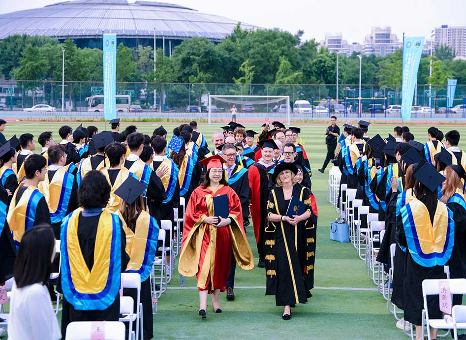 Graduates in academic gowns and caps seated in rows on a sports field, forming an aisle as faculty members in ceremonial robes process down the center during an outdoor graduation ceremony.
