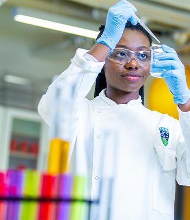 Student in a white lab coat using a pipette to transfer liquid in a laboratory setting.