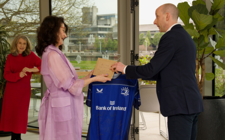 Man in dark suit presenting an envelope to dark haired woman in purple outfit