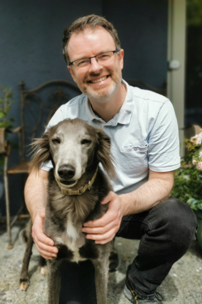 Mark McCorry smiling and kneeling outdoors beside a long-haired greyhound, with one hand gently resting on the dog’s chest.