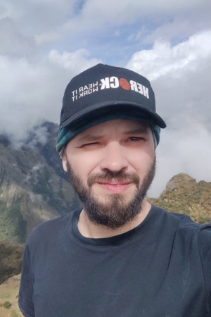Osvaldo Bogado Pascottini wearing a black T-shirt and a black cap that reads “HERDCK Learn It Work It” stands outdoors on a mountain with clouds and rugged peaks in the background.