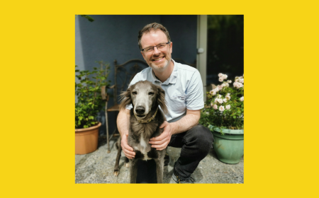 Mark McCorry kneeling beside a greyhound and smiling, surrounded by potted plants, with a bright yellow border framing the image.
