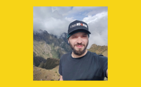Osvaldo Bogado Pascottini with a beard wearing a black T-shirt and black cap stands on a mountain trail with misty peaks behind him. The image is framed by a bright yellow background.